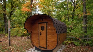 Una cabaña de madera con techo curvo, una puerta y un ventanal pequeño, rodeada de árboles y un camino de grava.