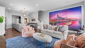A modern living room with a large cityscape painting, a kitchen, and a staircase. - Above the Gulch • Rooftop Fire Pit • Skyline Views (Nashville)