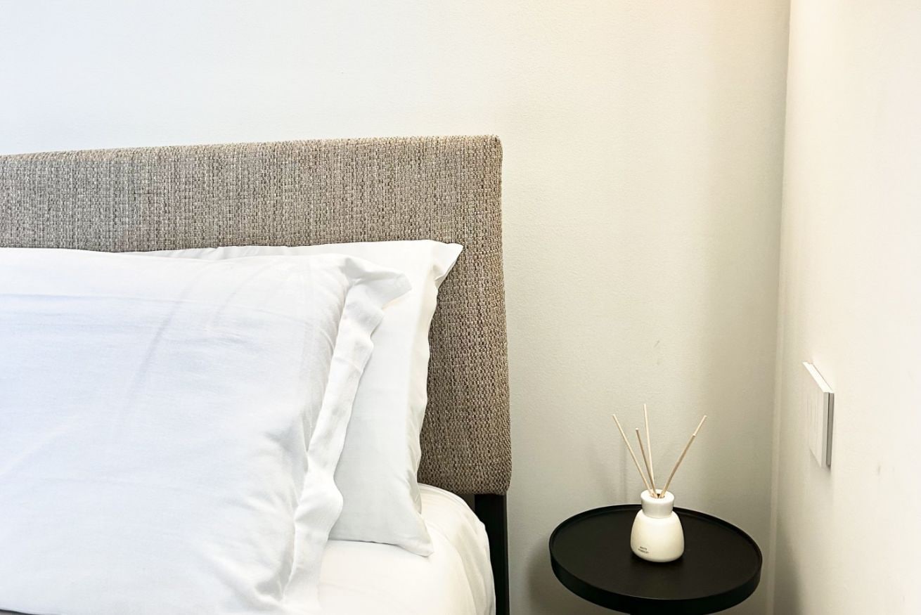 A bed with white bedding, a textured headboard, and a small table with a white vase and reed diffuser.