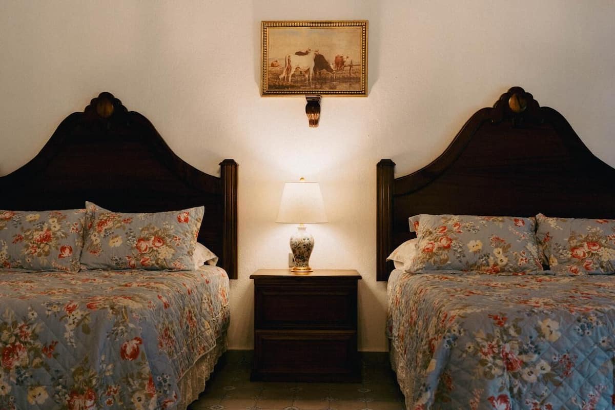 Une chambre avec deux lits, chacun agrémenté d’une literie à motifs floraux, une table de chevet avec une lampe et un tableau au mur.