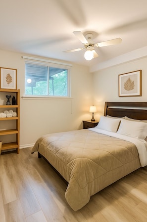 A bedroom with a bed, a ceiling fan, a bedside table with a lamp, a window with blinds, and a shelf with decorative items.