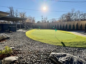 A well-maintained outdoor area with a curved green lawn, a wooden fence, and a covered structure. - Family Fun Hub • Games, Hot Tub, Putting Greens (Centennial)