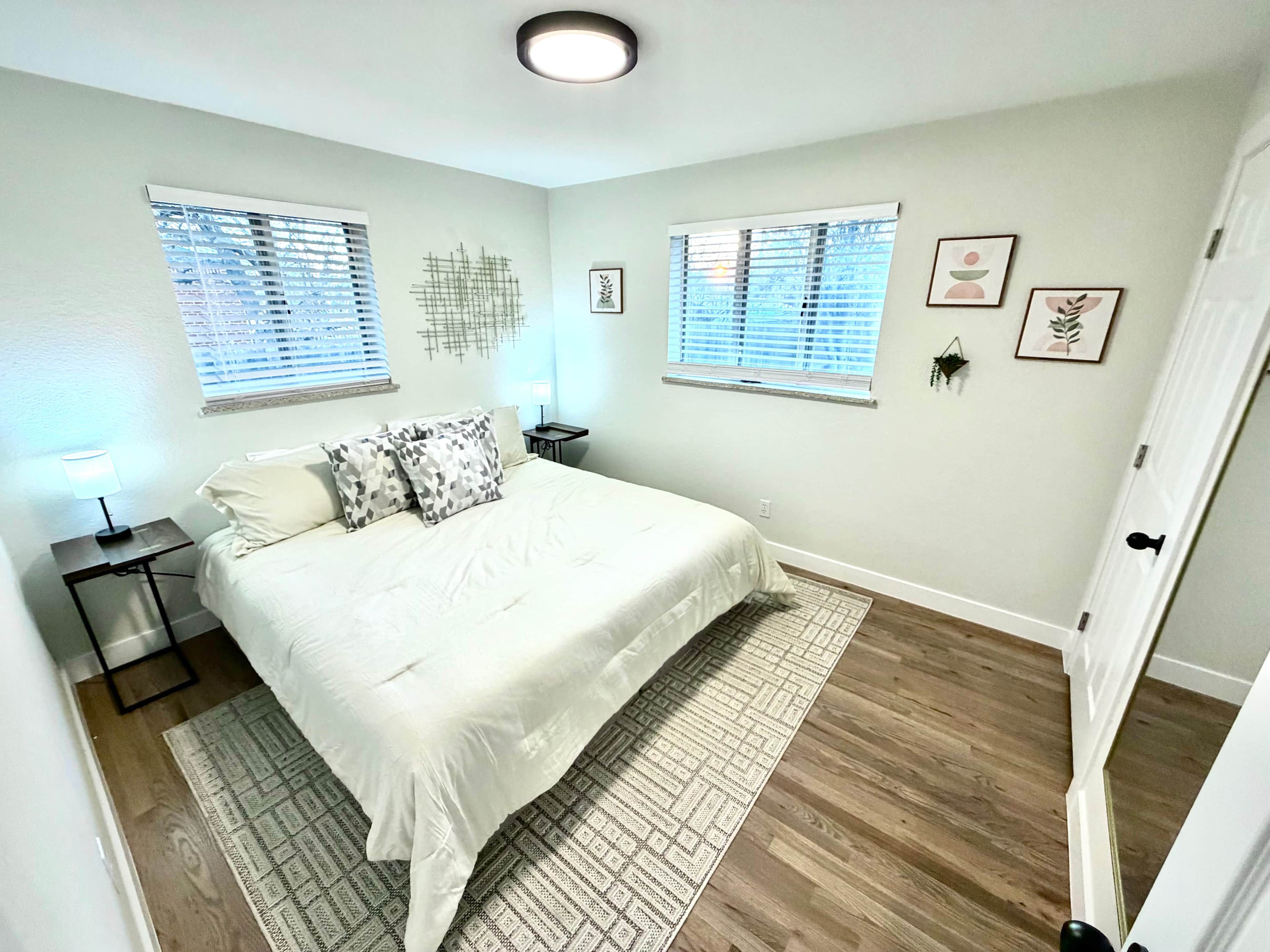 A bedroom with a bed, two bedside tables, a nightstand, a window with blinds, a wall-mounted artwork, and a door.