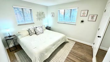 A bedroom with a bed, two bedside tables, a nightstand, a window with blinds, a wall-mounted artwork, and a door.
