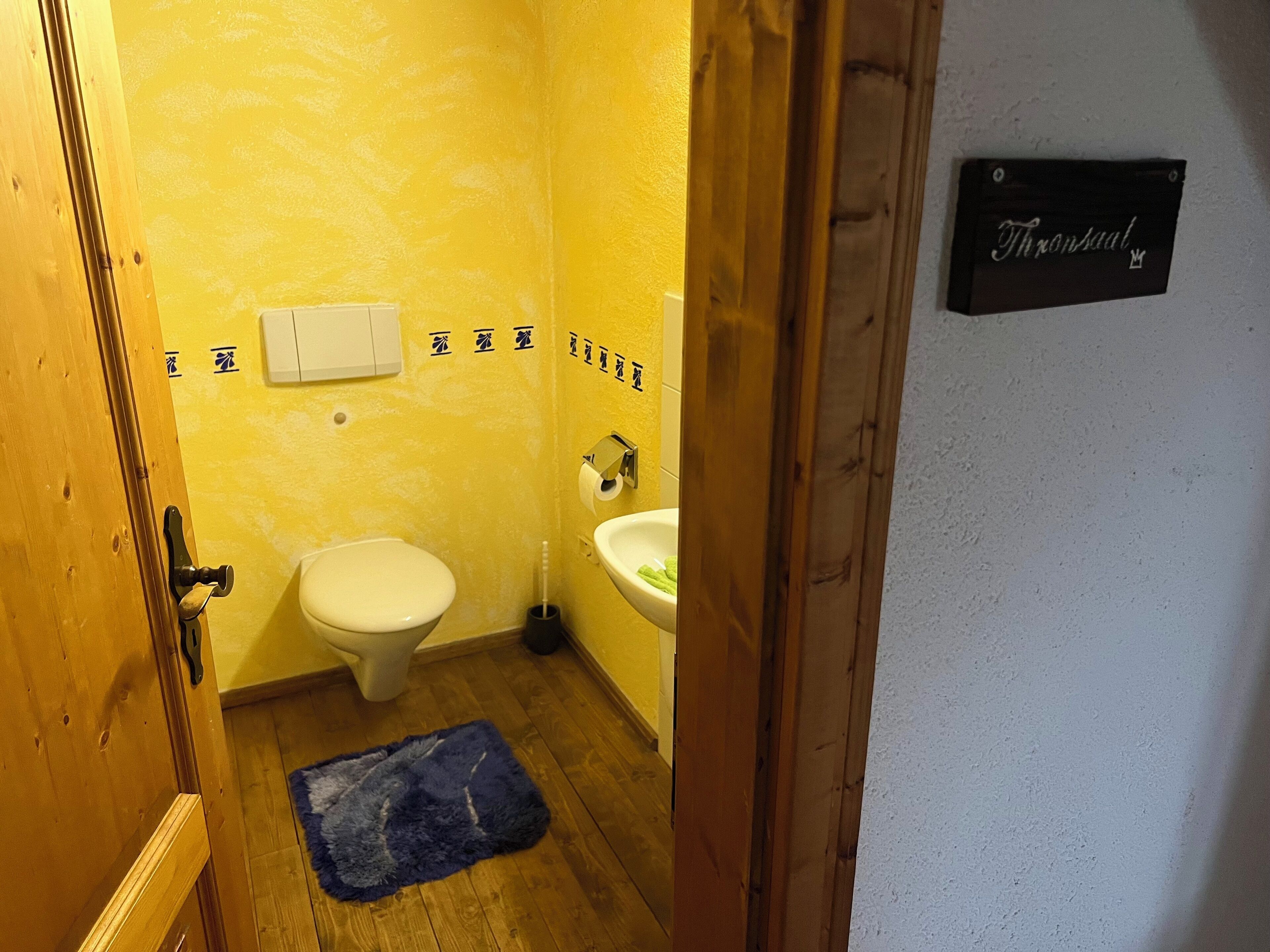 Ein kleines Badezimmer mit Toilette, Waschbecken und einer Holztür. An der Wand ist ein Schild mit der Aufschrift "Thronsaal".