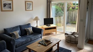 Een woonkamer met een blauwe bank, een houten salontafel, een televisie op een houten dressoir en een grote schuifpui die uitkomt op een tuin.