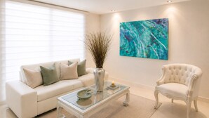 A living room with a white sofa, a glass coffee table, a white armchair, a vase with branches, and a large abstract painting on the wall. - Neos Apartment – Near the Sea by SOHO – 303A (Santa Marta)