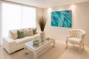 A living room with a white sofa, a glass coffee table, a white armchair, a vase with branches, and a large abstract painting on the wall. - Neos Apartment – Near the Sea by SOHO – 303A (Santa Marta)