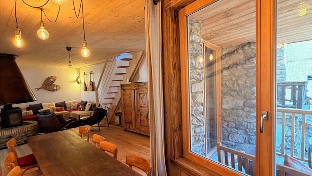 A wooden interior with a long dining table, multiple chairs, a fireplace, and a large window with a stone wall view. - Spacious 10-person chalet (150 m², 4 bedrooms) at the foot of the slopes. (Vars)