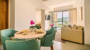 Un salon moderne avec une table à manger ronde, des chaises recouvertes d’un tissu vert, un canapé beige et une grande fenêtre donnant sur la ville.