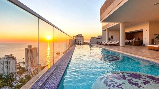 Une piscine à débordement située sur le toit, offrant une vue imprenable sur le coucher de soleil sur l’océan, et entourée d’immeubles modernes.