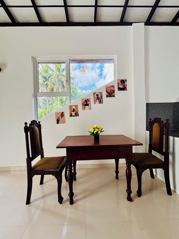 A dining area with a wooden table and chairs, a window with a view of greenery, and a wall decorated with pictures.