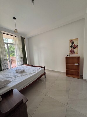 A bedroom with a bed, a wooden nightstand, a small wooden shelf, a ceiling fan, and a window with curtains.