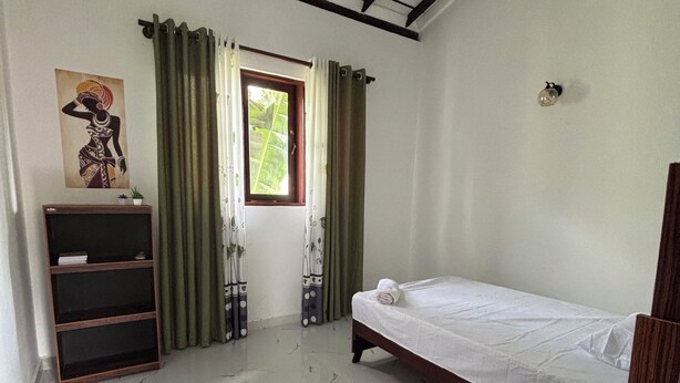 Un dormitorio con cama, estante, ventana con cortinas, luz de techo y un despertador en la pared.