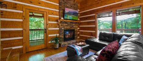 A log cabin interior with a stone fireplace, a black leather sofa, and a wooden deck visible through a glass door.