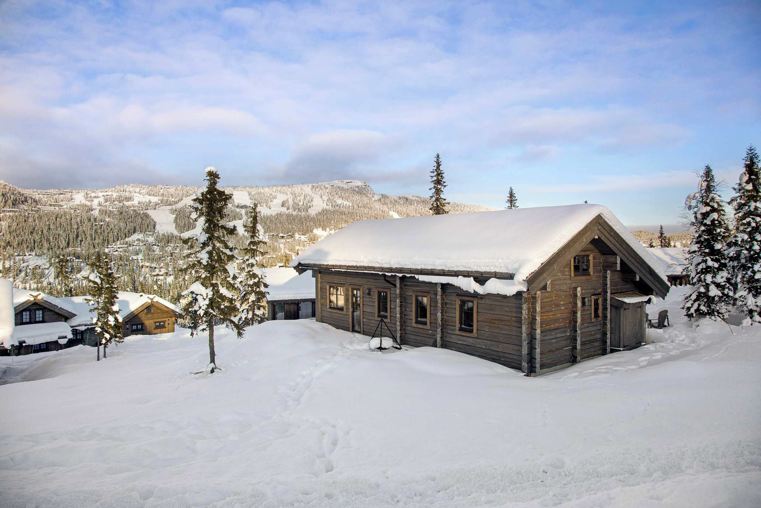 A wooden cabin with a snow-covered roof, surrounded by snow-covered trees and a mountainous backdrop.
