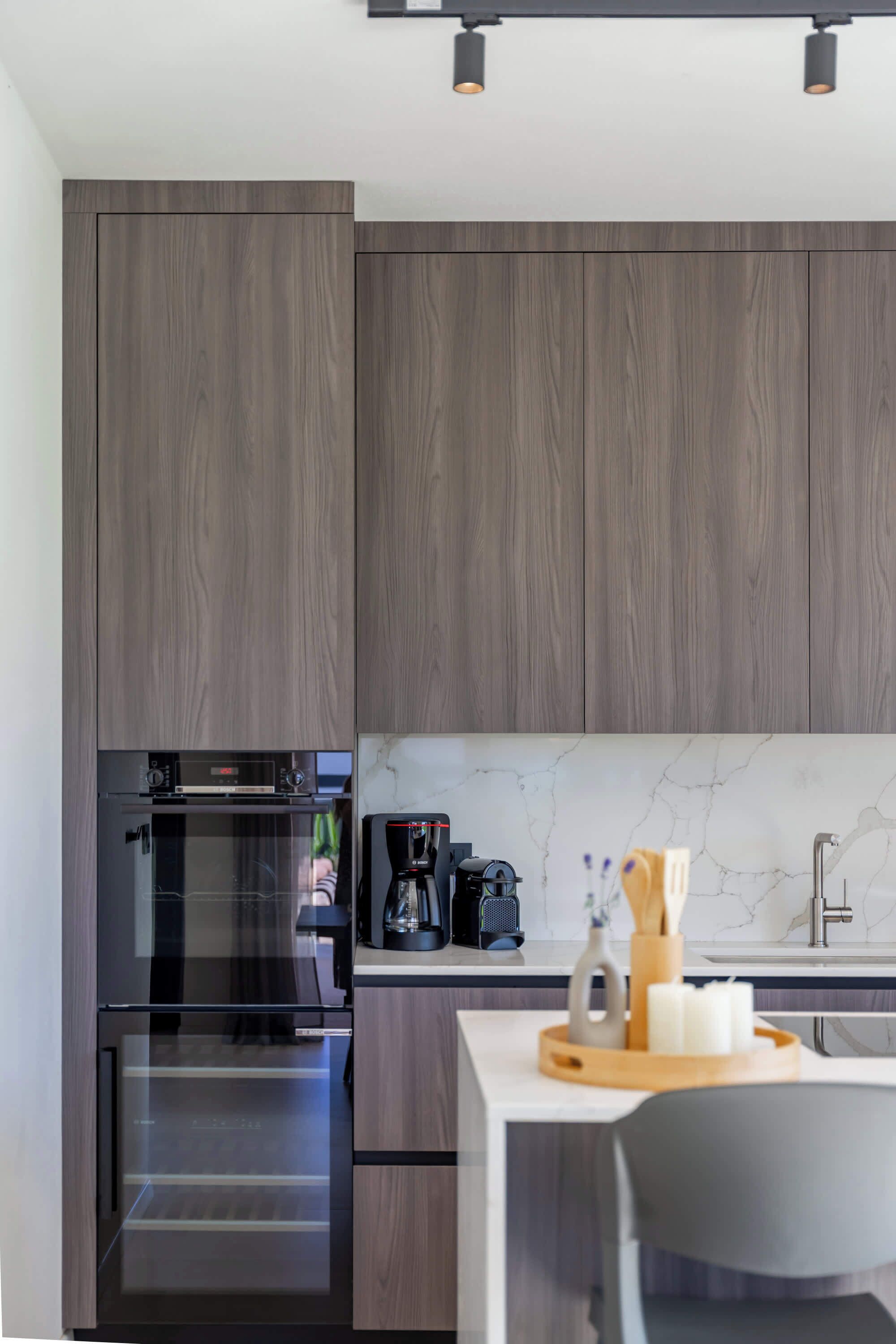 A modern kitchen with dark wood cabinets, a built-in oven, a coffee maker, and a marble countertop.