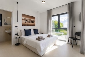 Ein modernes Schlafzimmer mit einem großen Bett, einem Badezimmer mit einem Waschbecken, einem Bild an der Wand und Blick durch das Fenster.
