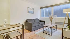Een moderne woonkamer met een grijze bank, een salontafel, een eettafel met stoelen en een groot raam met uitzicht op een bakstenen gebouw.
