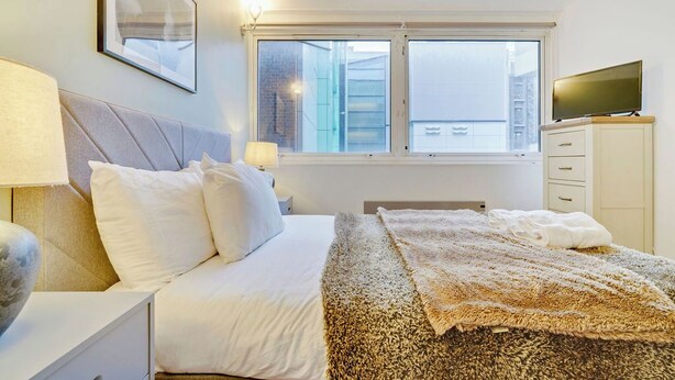 Een slaapkamer met een bed, nachtkastjes, een televisie en een raam met uitzicht op gebouwen.