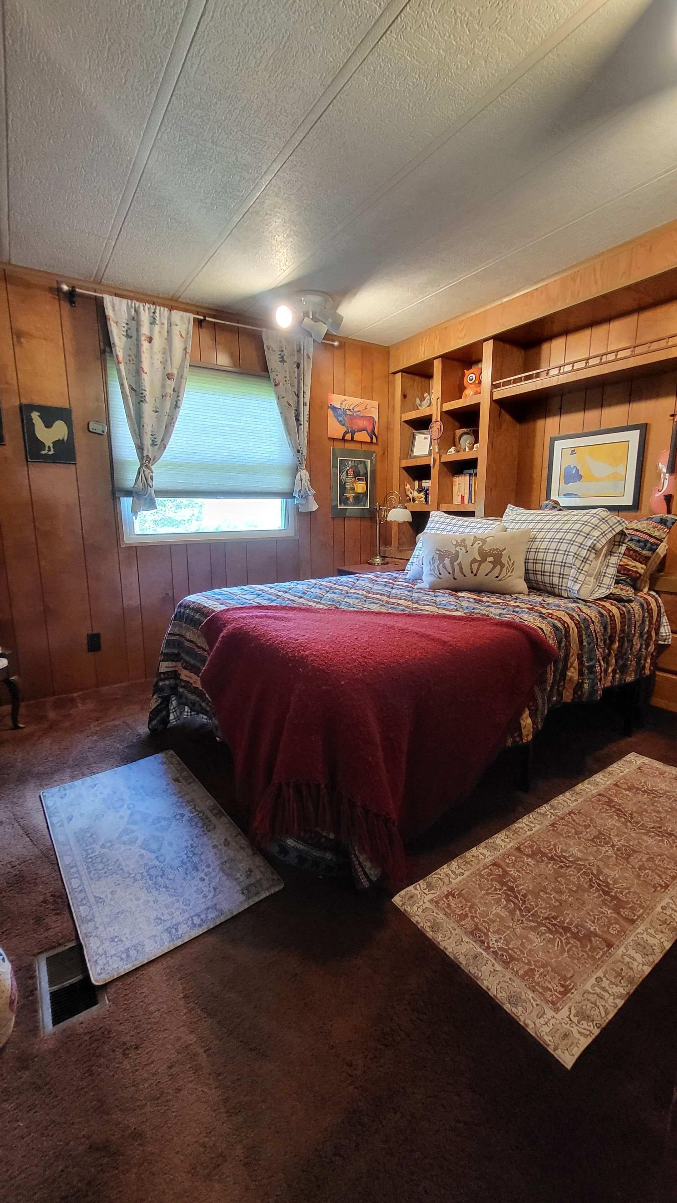 A bedroom with a bed, a window with curtains, a wooden headboard, a shelf with decorative items, and a rug.