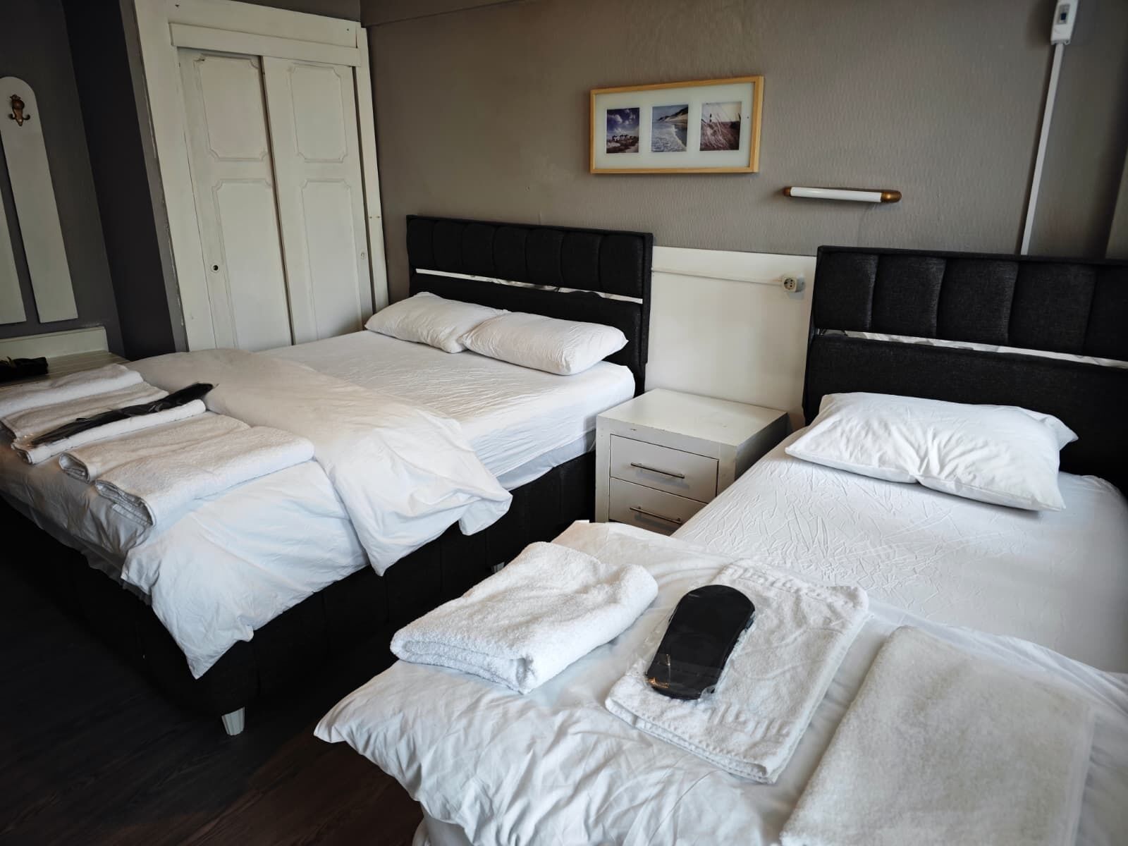 A hotel room with three beds, each with white linens and pillows. There is a small bedside table with a lamp and a drawer. The room has a neutral color scheme with a framed picture on the wall.