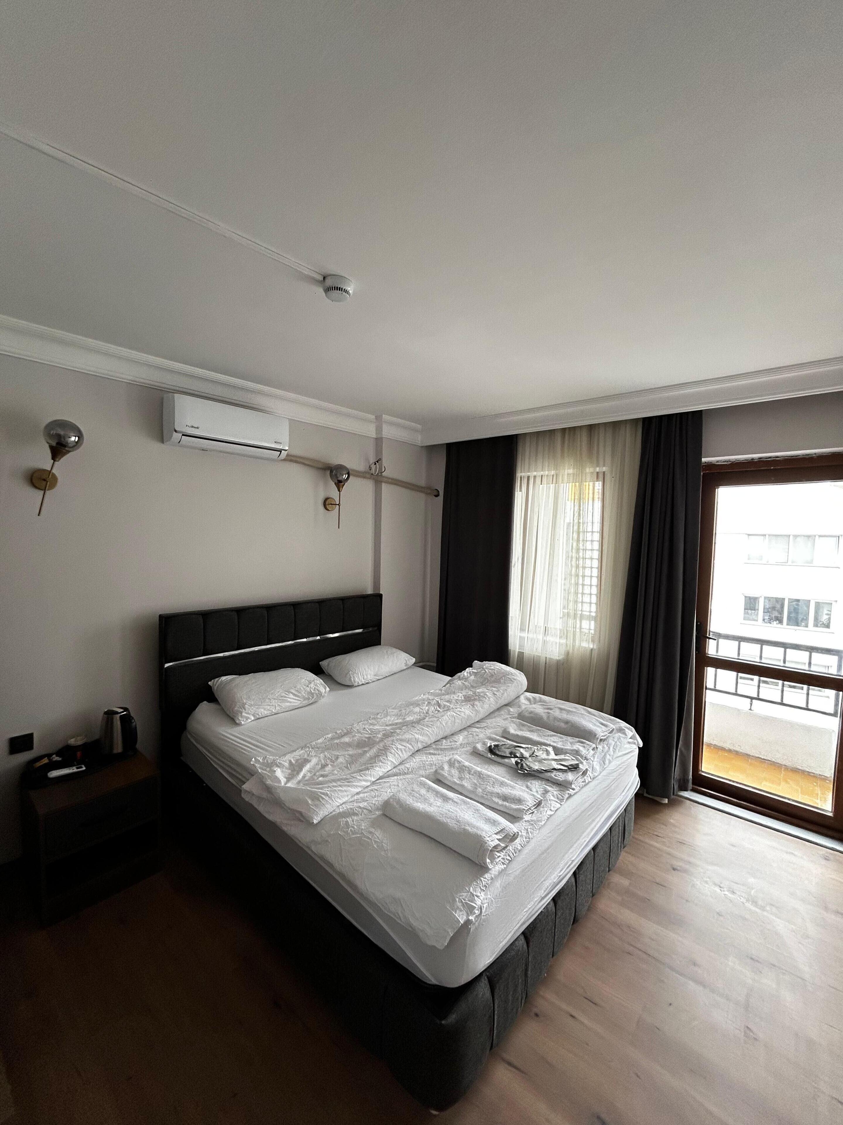 Una habitación de hotel con una cama grande, un equipo de aire acondicionado, una mesita de noche y una ventana con cortinas.