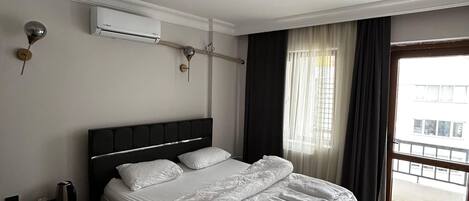 Una habitación de hotel con una cama grande, un equipo de aire acondicionado, una mesita de noche y una ventana con cortinas.
