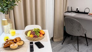 Une chambre d’hôtel avec une table à manger dressée, agrémentée de croissants, d’un panier de fruits et d’un verre de jus d’orange. Un téléviseur est fixé au mur et il y a un bureau avec un ordinateur.