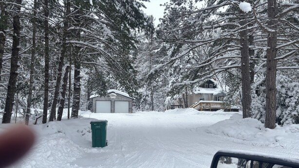 Una strada residenziale innevata, con un garage e un bidone della spazzatura verde.