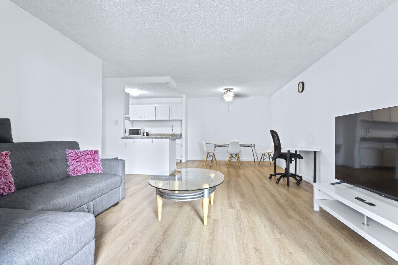A modern living room with a grey sofa, a glass coffee table, a dining area with a white table and chairs, and a TV on a white stand.