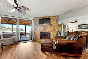 A living room with a stone fireplace, leather furniture, a ceiling fan, and a view through large glass doors.