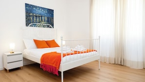 A neatly made bed with white and orange bedding, a white nightstand with a lamp, and a painting of a night scene on the wall. - Dimora Aniene - Two Bedroom Apartment, Sleeps 4 (Rome)