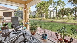 A covered patio with wicker furniture, potted plants, and a view of a residential area with trees and buildings.