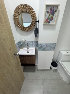 A bathroom with a wooden door, a white sink with a marble countertop, a round mirror with a woven frame, a black towel, a small trash bin, and a toilet. - Mussi House Resort in beautiful Sopetrán with cool AC and private pool (Sopetrán)