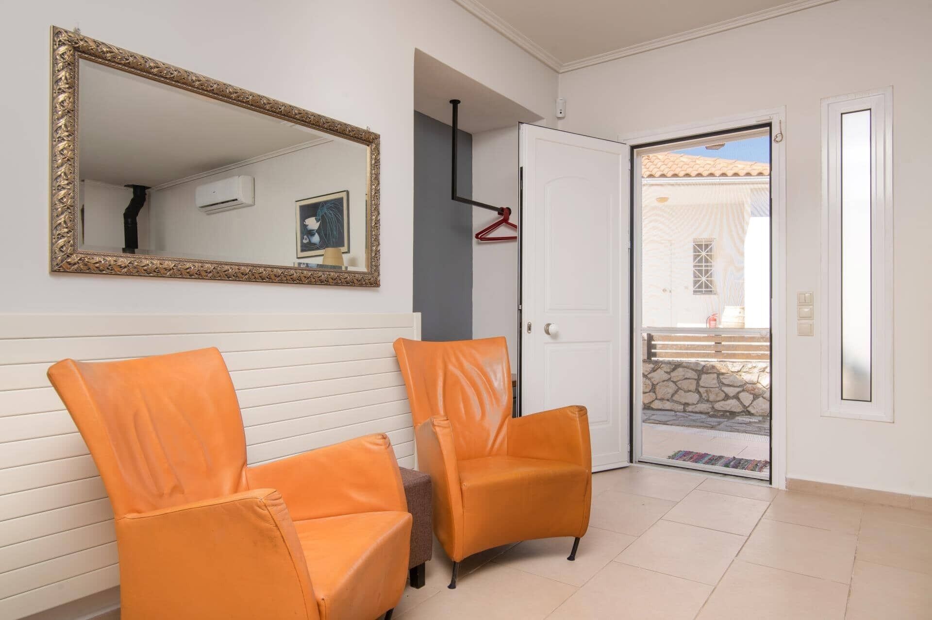 Una habitación con dos sillas anaranjadas, un espejo grande y una puerta blanca que da al exterior.