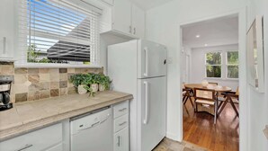 Eine Küche mit weißen Schränken, einem weißen Kühlschrank, einer gefliesten Spritzschutzleiste, einer Kaffeemaschine und einem Essbereich mit Holztisch und Stühlen.