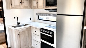 Una cucina compatta con lavandino, piano cottura, microonde e frigorifero, caratterizzata da mobili in legno chiaro e un piano di lavoro bianco.