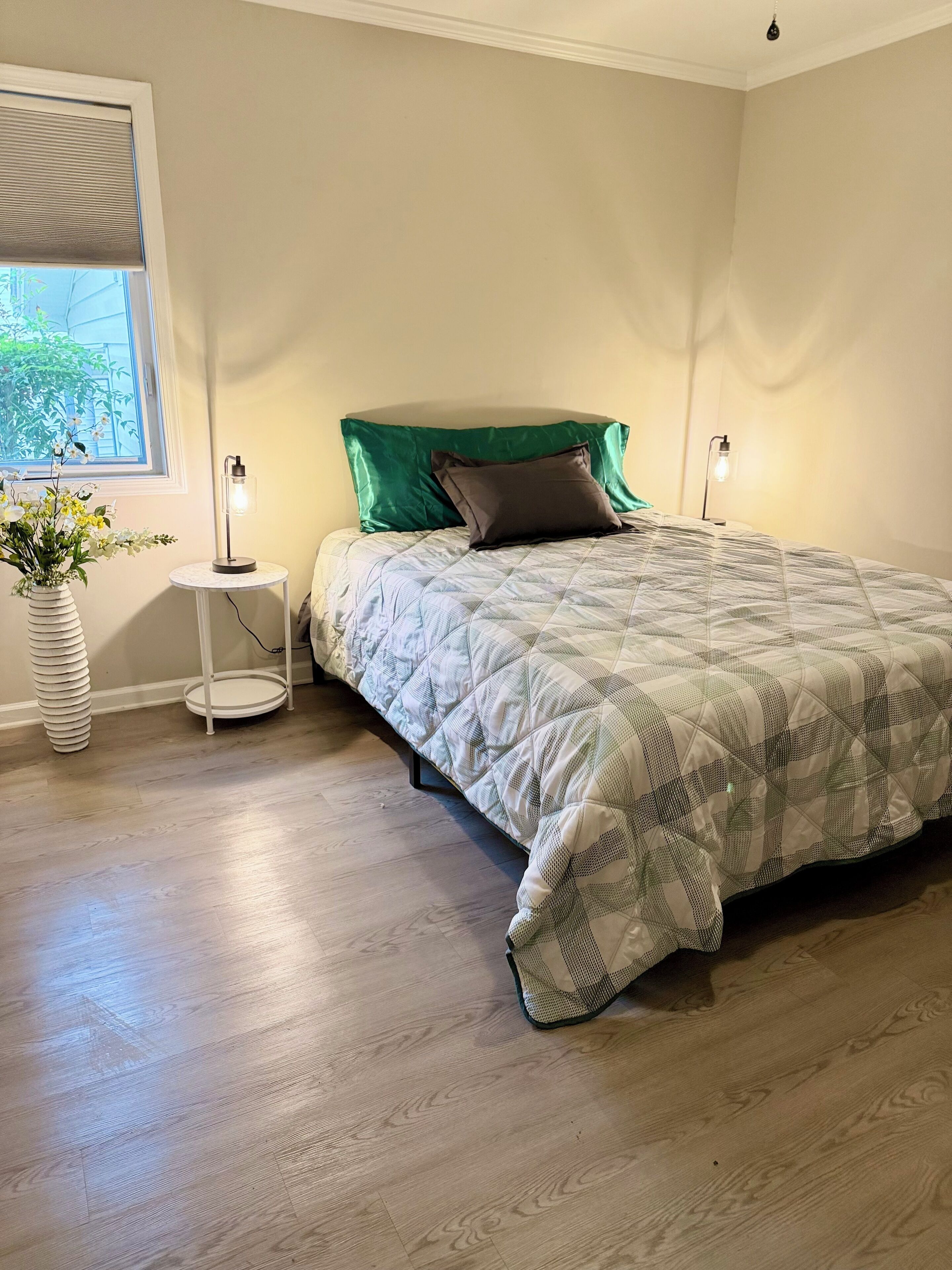 A bedroom with a bed, a window with blinds, a bedside table with a lamp, and a floor lamp.