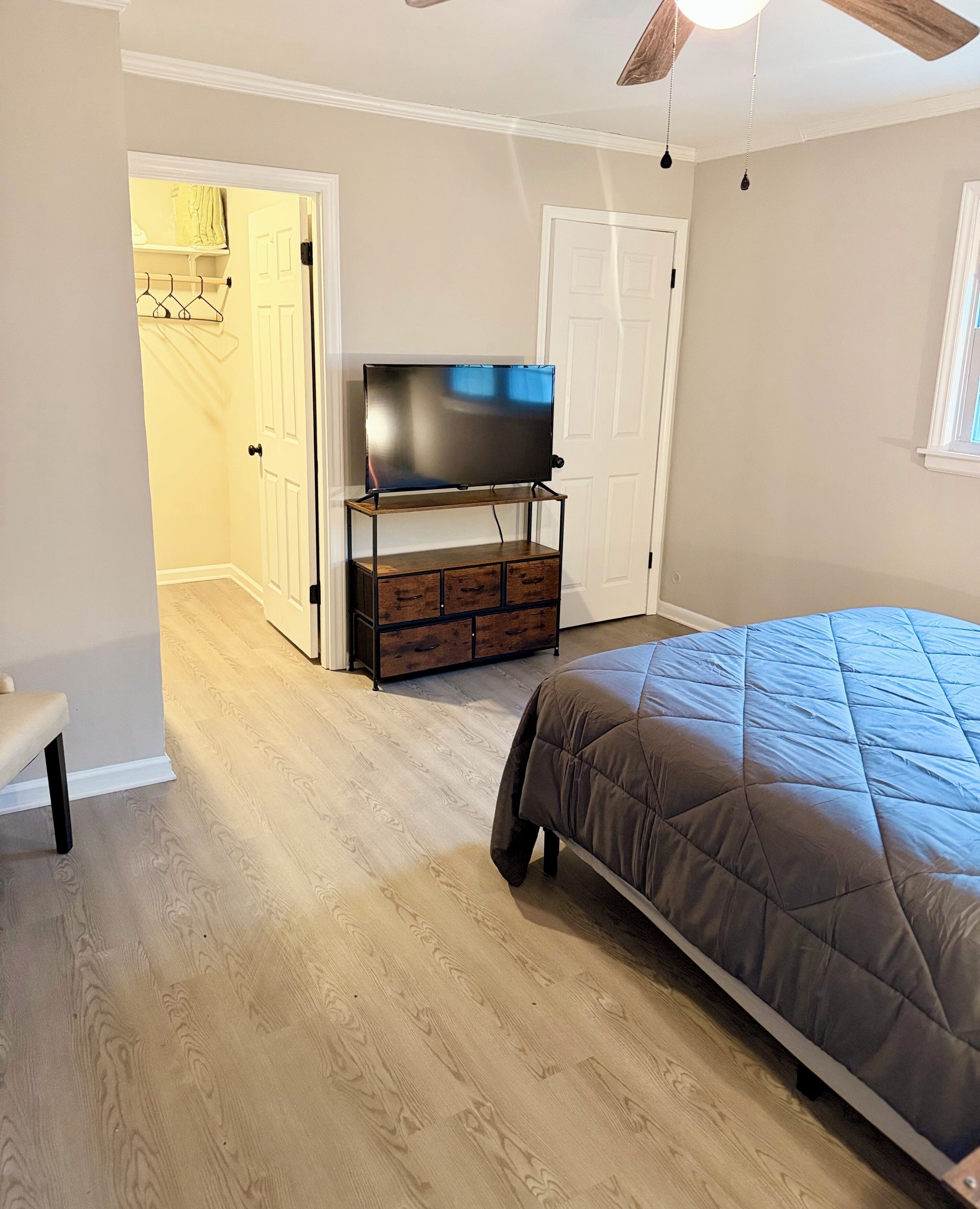 A bedroom with a bed, a TV on a wooden stand, a bench, and a door leading to another room.