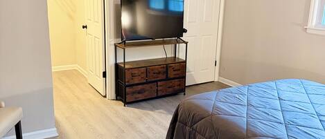 A bedroom with a bed, a TV on a wooden stand, a bench, and a door leading to another room.