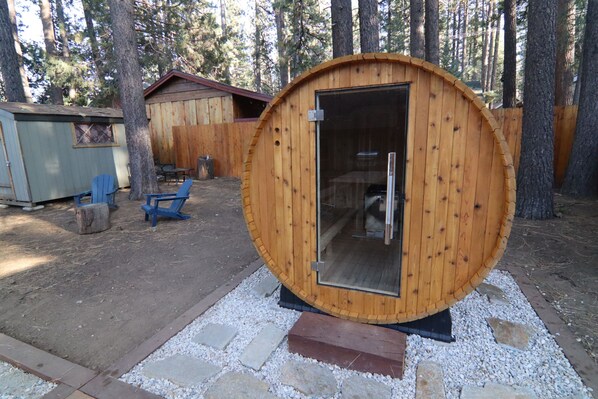 A wooden cabin with a glass door, a small outdoor seating area, and a surrounding forest.