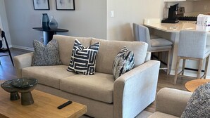 Una sala de estar con un sofá color beige, dos almohadones estampados, una mesa de centro de madera y un control remoto.