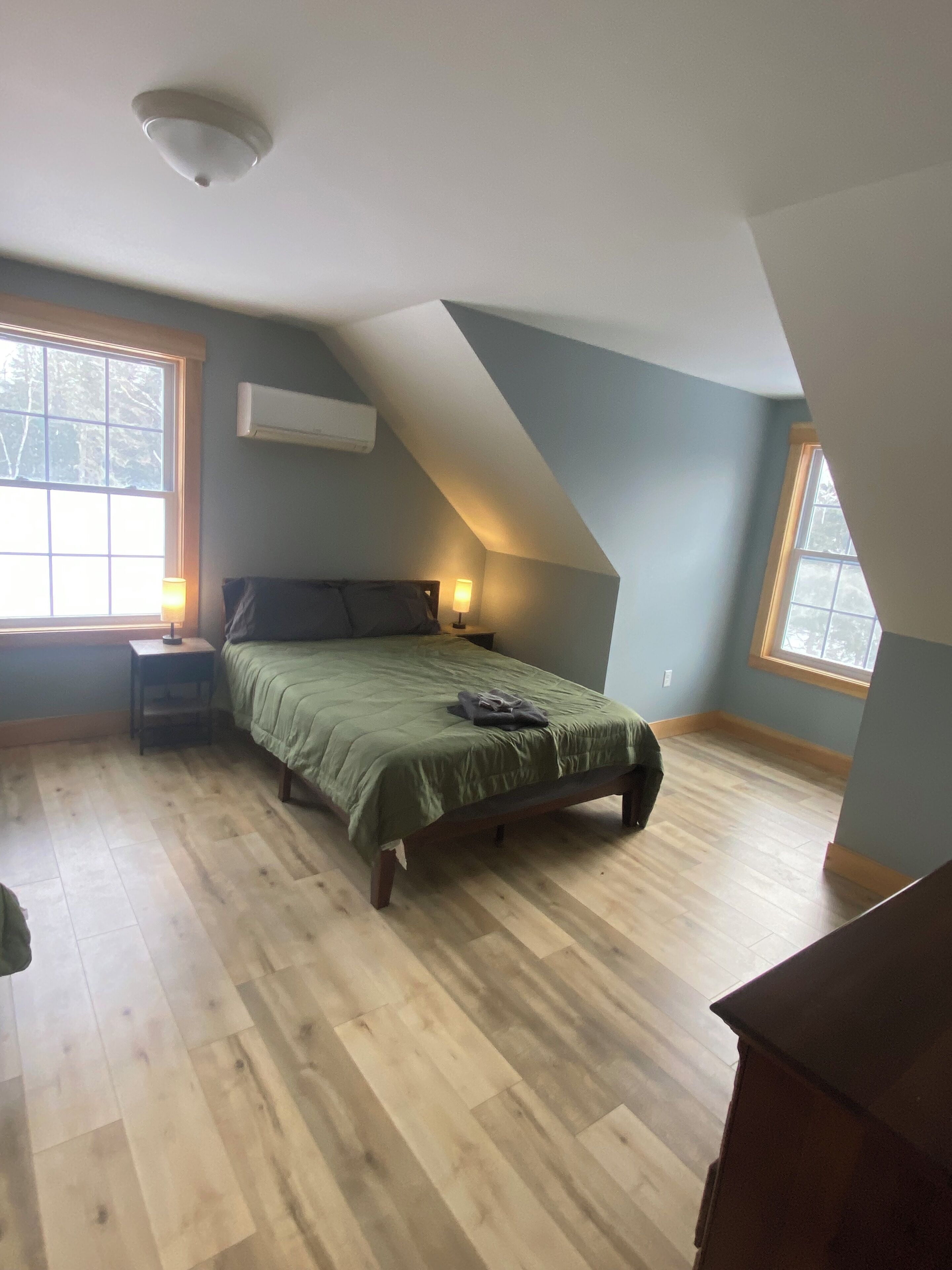 A bedroom with a sloped ceiling, a bed with a green bedspread, two bedside lamps, and a window with a view of trees.