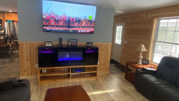 A living room with a flat-screen TV mounted on the wall, wooden flooring, a sofa, a coffee table, and a small side table. - Beautiful 6-bedroom house in Rangeley perfect for friends and family getaways. (Rangeley)