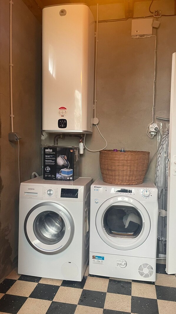 Deux machines à laver à chargement frontal, un chauffe-eau et un panier en osier dans une buanderie dont le sol est à carreaux.