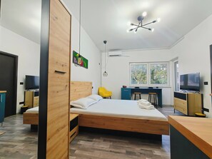 Une chambre moderne avec un lit en bois, un bureau avec une chaise, une télévision et une fenêtre donnant sur des arbres.