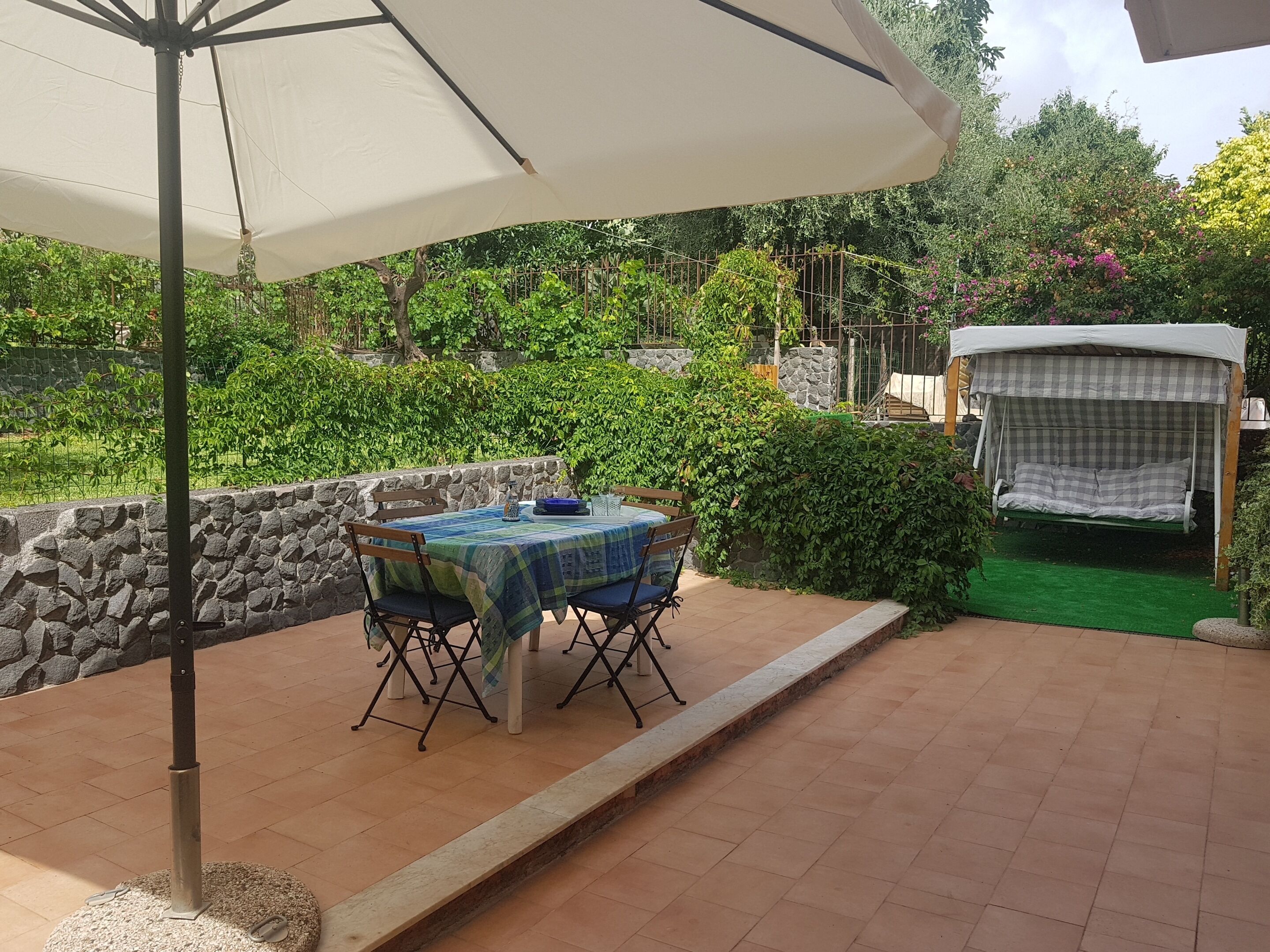 An outdoor patio with a table and chairs under a canopy, surrounded by greenery and a stone wall.