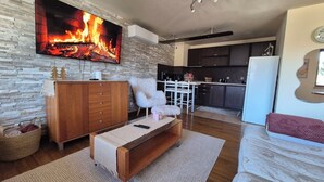 A cozy living room with a fireplace, a wooden cabinet, a fluffy chair, and a kitchen in the background. - 3 bedroom Penthouse only 700 meters from Ski Lift (Bansko)