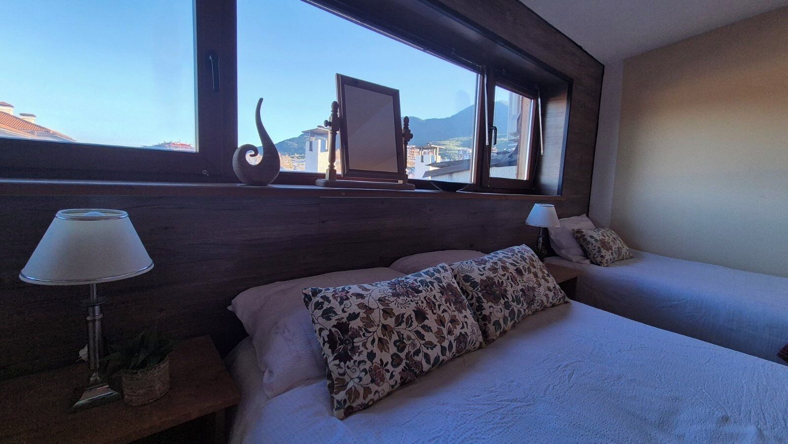 A bedroom with two beds, a window with a view of mountains, and a lamp on the nightstand.